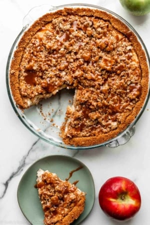 apple pie cheesecake topped with caramel sauce with 1 slice missing that's placed on green plate.