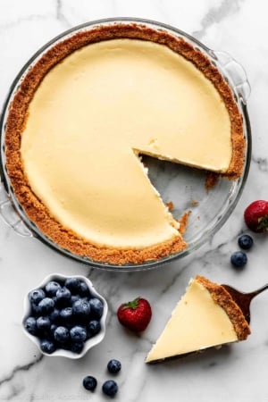 cheesecake pie in glass pan with slice taken out and blueberries and strawberries scattered next to the dish.