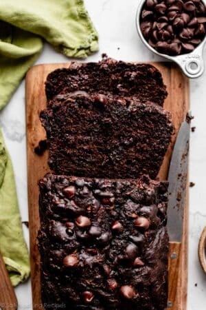 loaf of chocolate zucchini bread with chocolate chips cut with 2 slices on wooden cutting board.