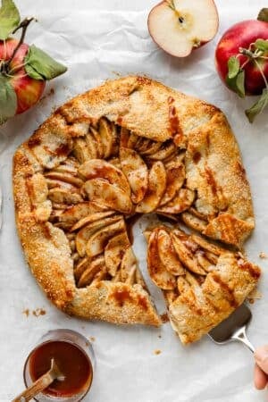 apple galette with caramel sauce on white parchment paper with apples and jar of caramel.