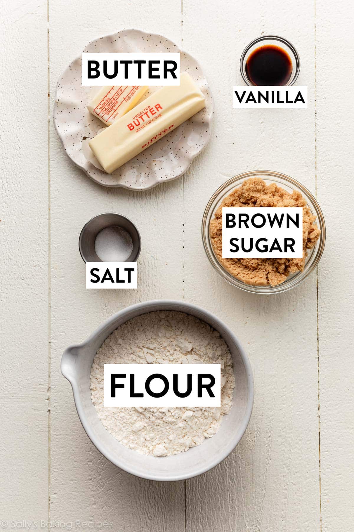 ingredients on white wooden backdrop including flour, butter, vanilla, and salt.