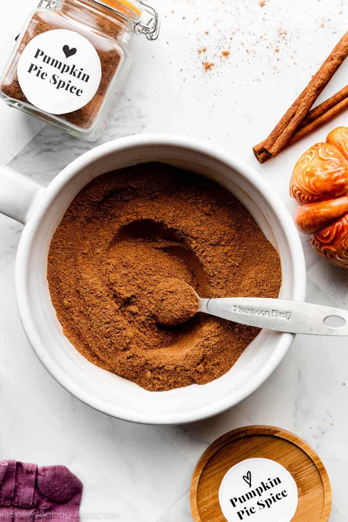 homemade pumpkin pie spice in white bowl with labeled container and cinnamon sticks next to it.