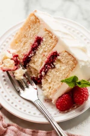 3 layer white vanilla cake slice on white plate with vanilla frosting and raspberry cake filling in the layers.