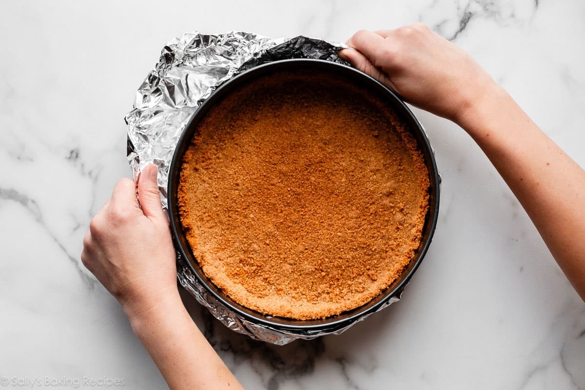 hands wrapping outside of springform pan with graham cracker crust with aluminum foil.